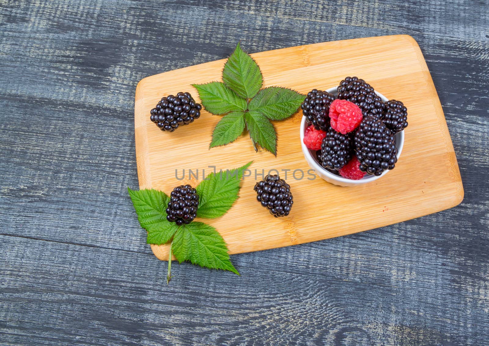 ripe blackberry with leaves on a wooden cutting board in a white ceramic bowl on dark blue wooden background.