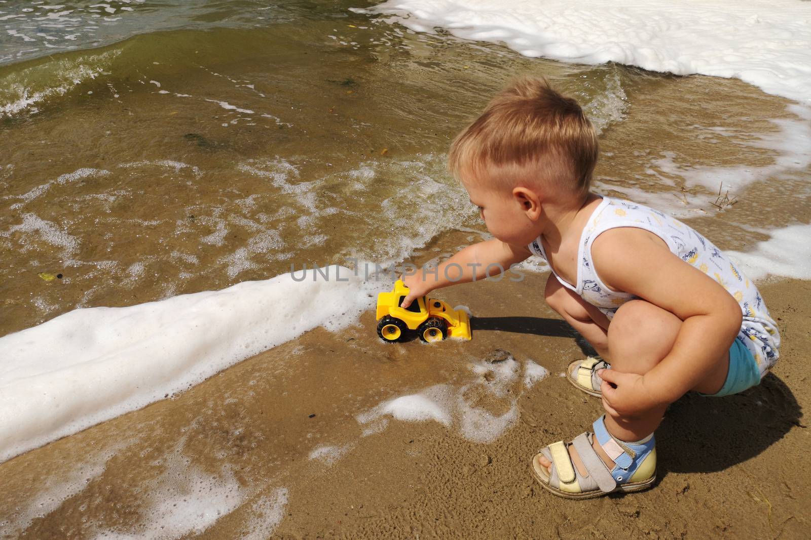 A cute little boy plays on the shore of the lake with a bright yellow toy car in the foam of the lake.Summer kids holiday concept. Rest on the shore.