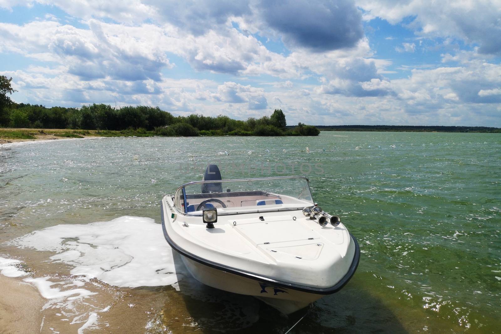 White motor boat motorboat stands near the shore of the lake against the background of blue sky with clouds.