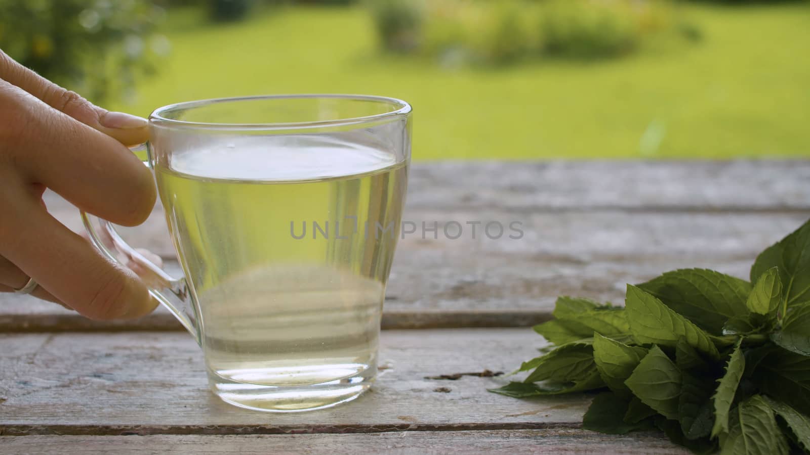 Close-up hand puts a transparent cup of mint tea or tisane on the old wooden table outdoors. A fresh mint on the table. Herbal medicine concept