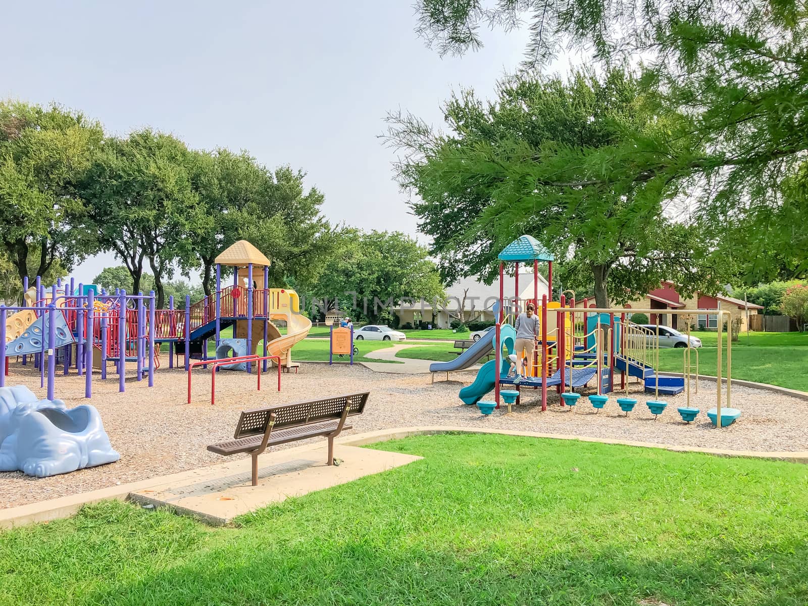 Empty metal bench at colorful playground near residential neighborhood in Richardson, Texas, America. Community facility surrounded by large oak trees, green grass lawn and single family houses