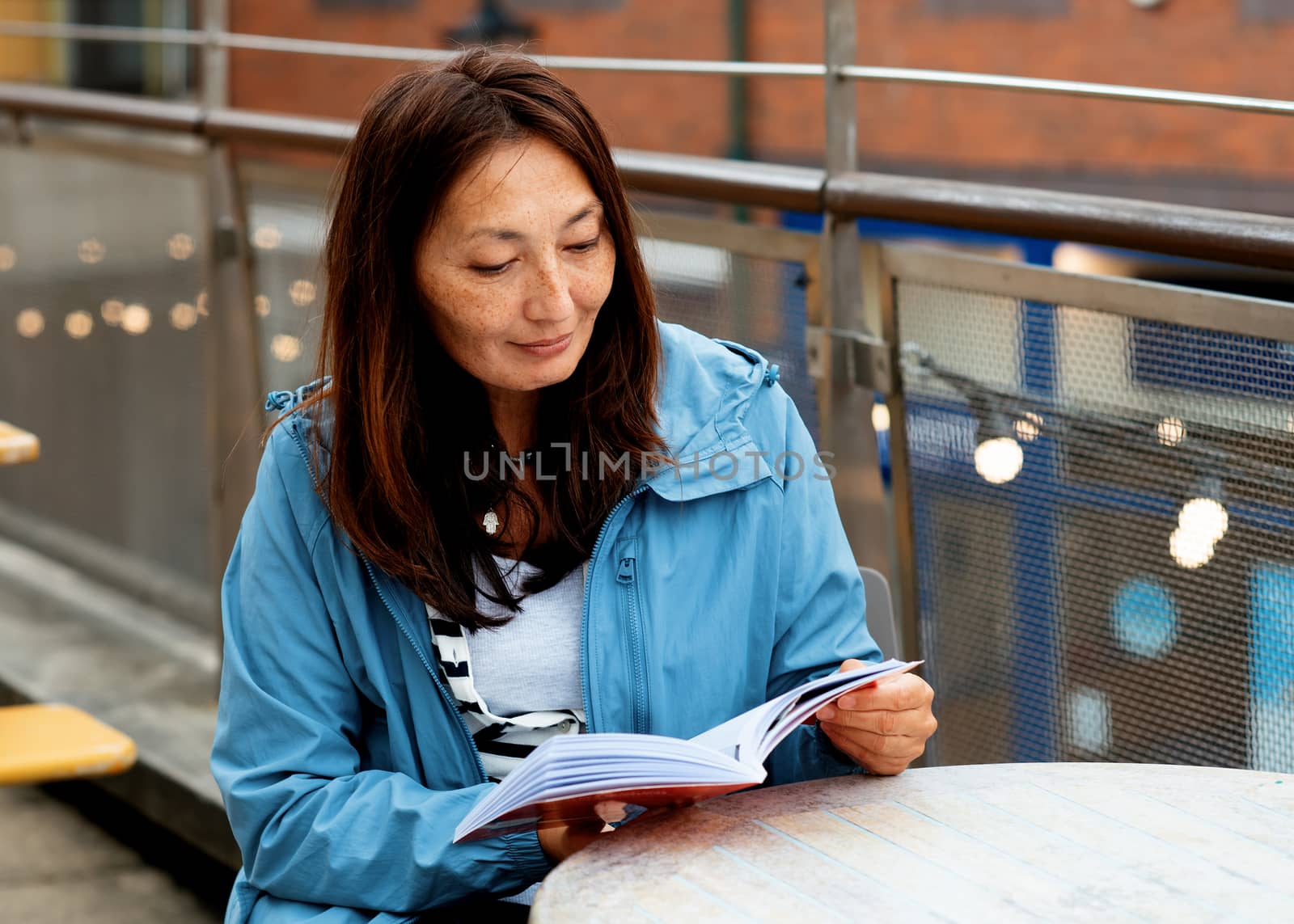 Asian woman in a blue coat sitting in the cafe in the street and reading a book