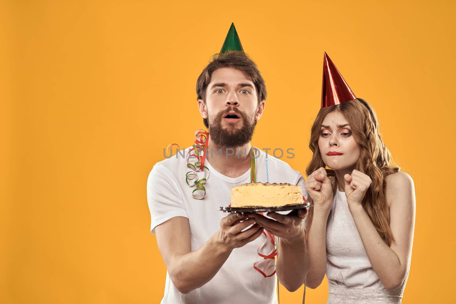 Happy man and woman in a cap celebrating a birthday on a yellow background with a cake in their hands. High quality photo