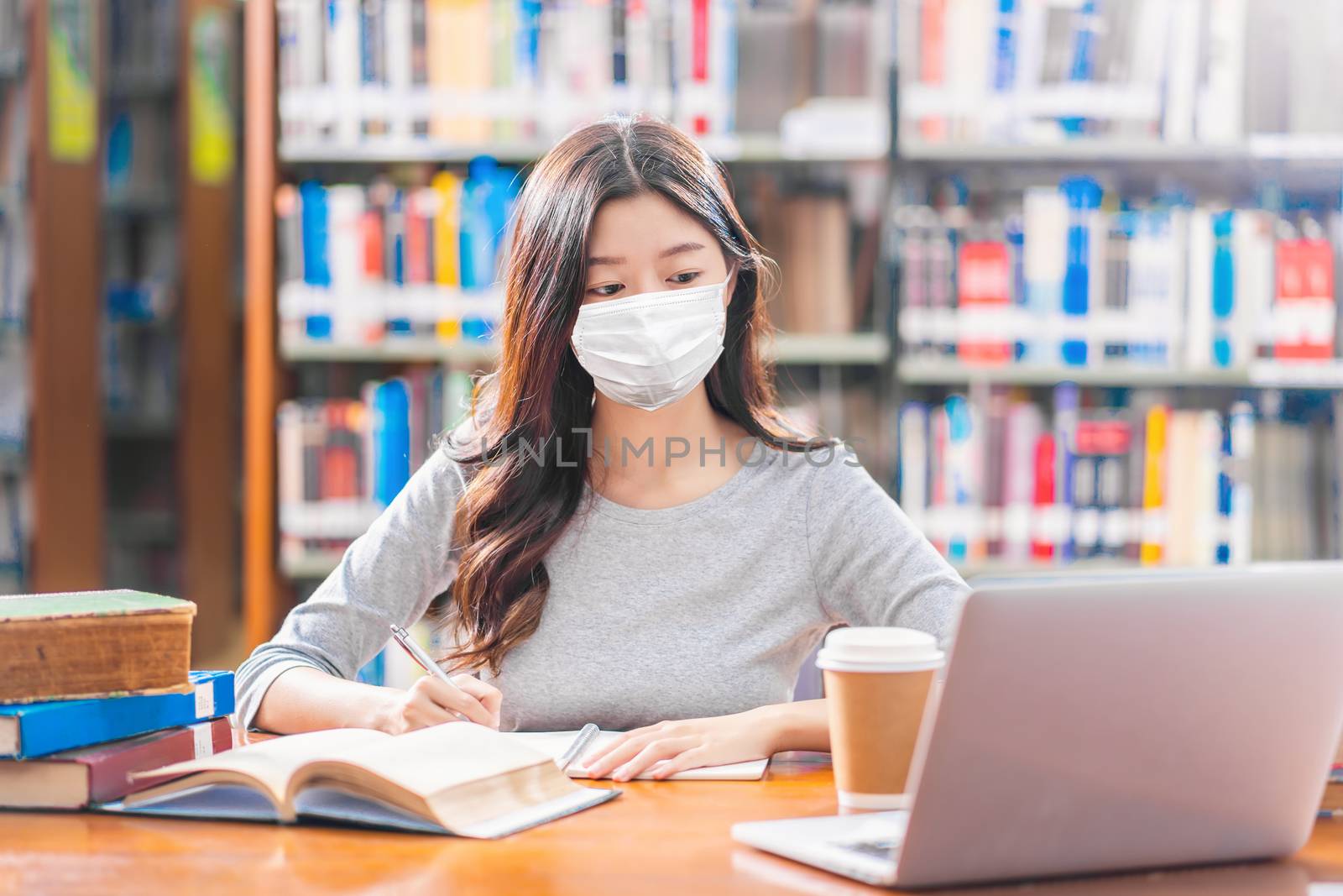 Asian young Student in casual suit wearing surgical mask and doing homework and using technology laptop in library of university or colleage with various book and stationary over book shelf background