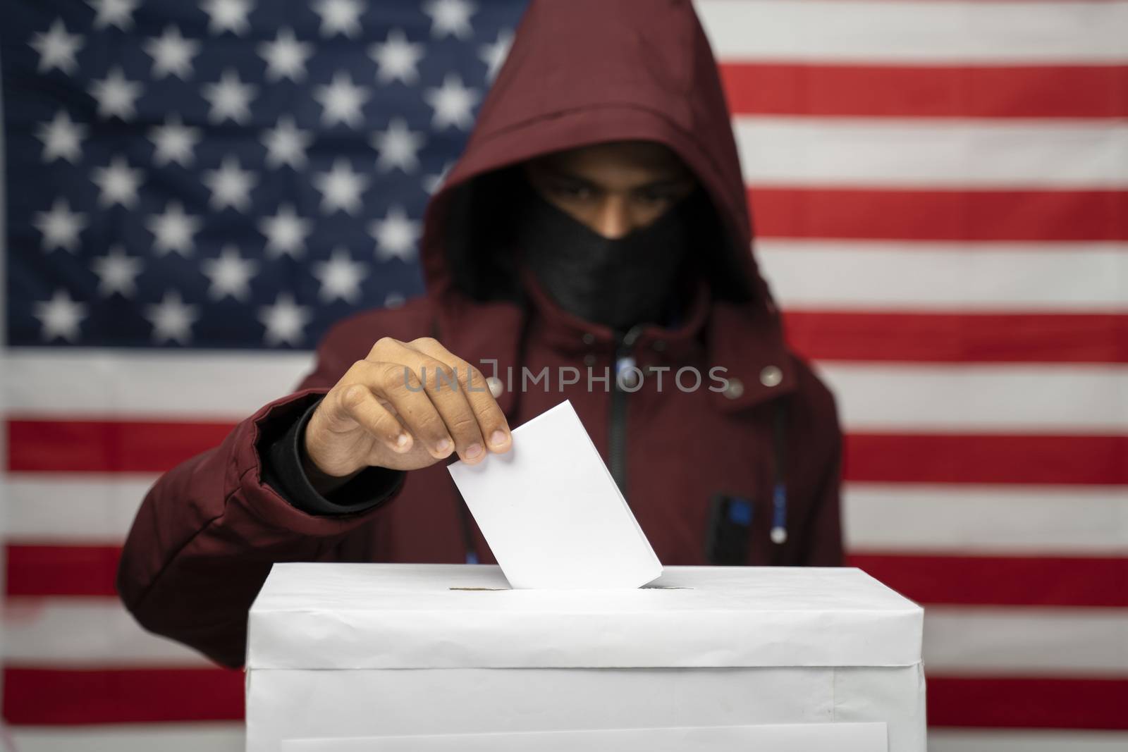 Man in hoodie with face covered casting Vote at polling booth with US falg as background - Concept of unkonwn voting or vote rigging in US elections by lakshmiprasad.maski@gmai.com