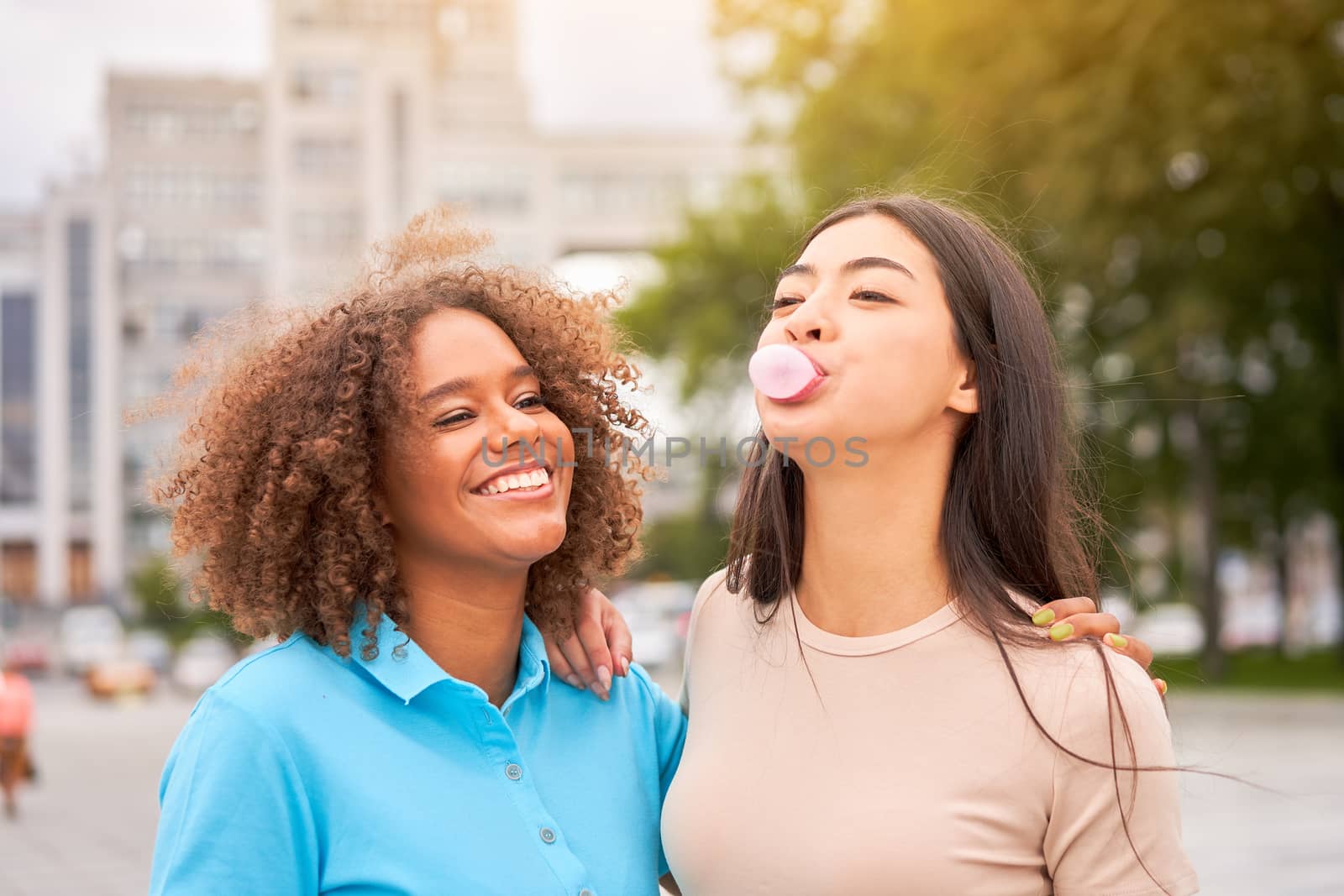 African american woman laughing while asian friend inflating bubble gum. Closeup face of multiethnic friends enjoying outdoor street. Girl laughing and blowing chewing gum with friend embracing her