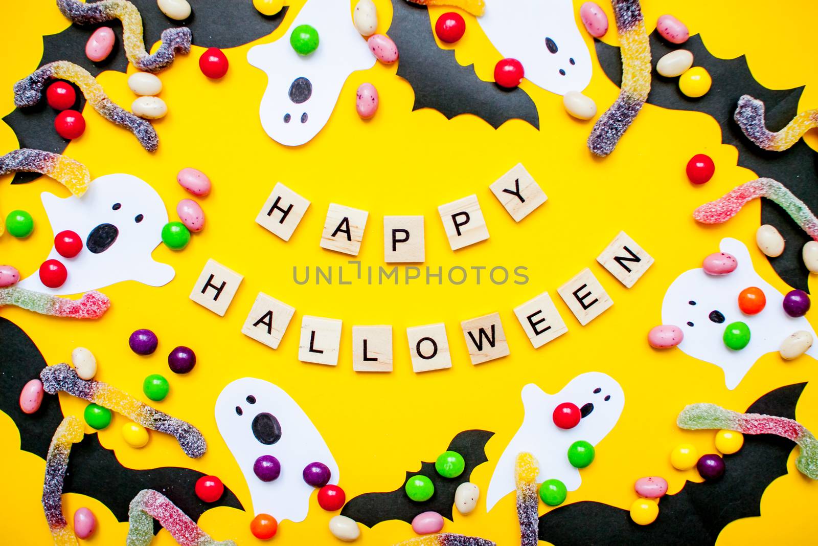 inscription from wooden blocks Happy Halloween and frame made of paper homemade bats and paper ghosts and multicolored candies and worms from gummy on a bright yellow background