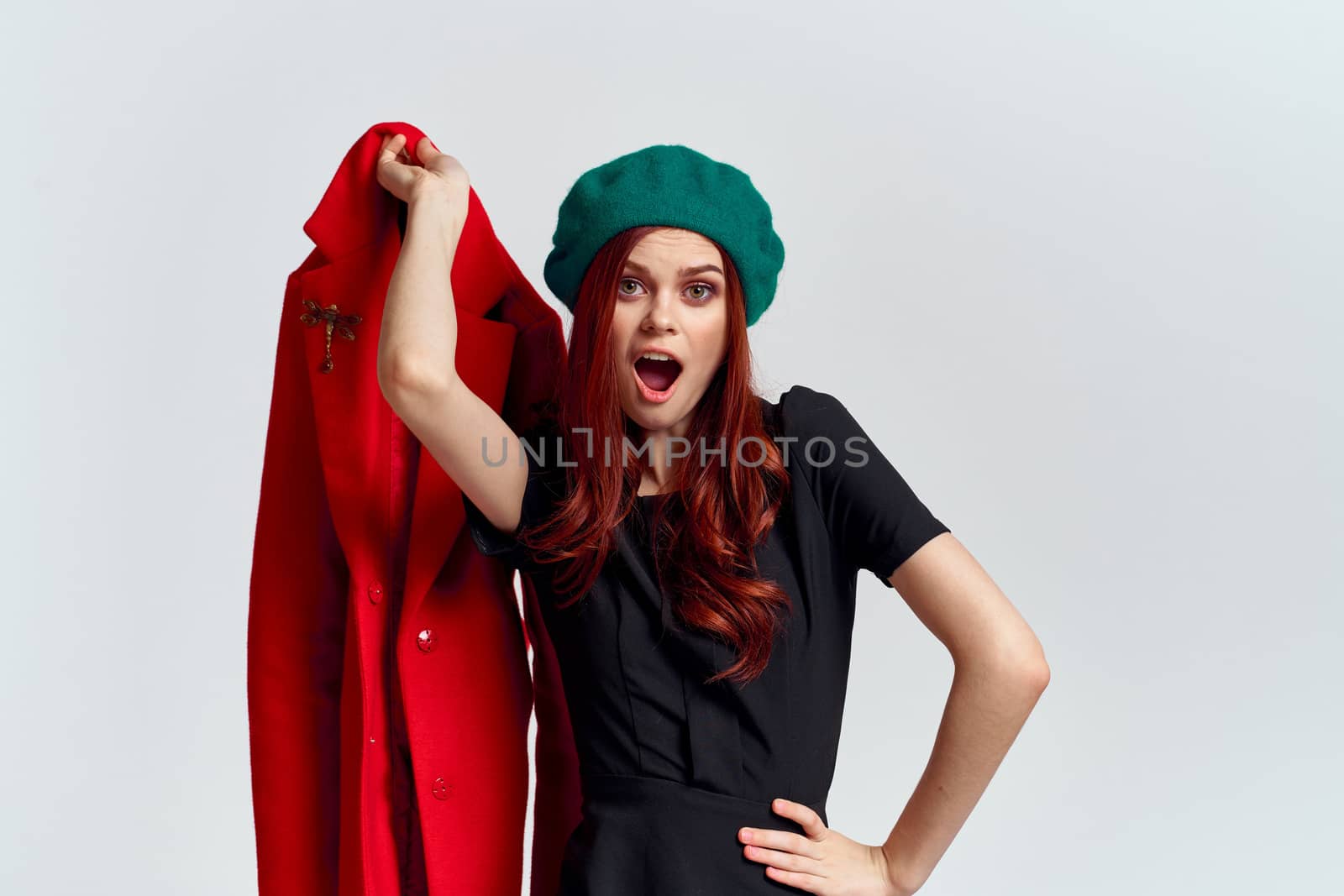A woman holds in her hand a red coat and a dark T-shirt Green hat cropped view of a light background by SHOTPRIME