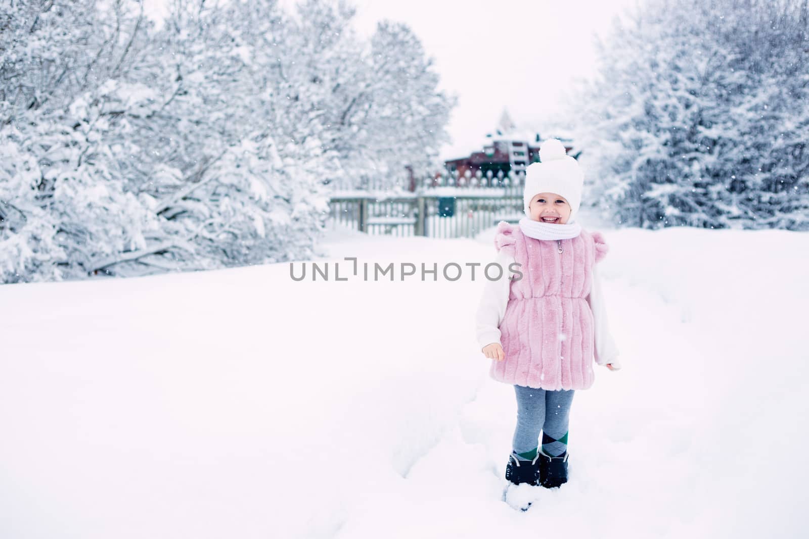.A little girl in a pink fur coat stands in the middle of a snow-covered park on a cloudy winter day