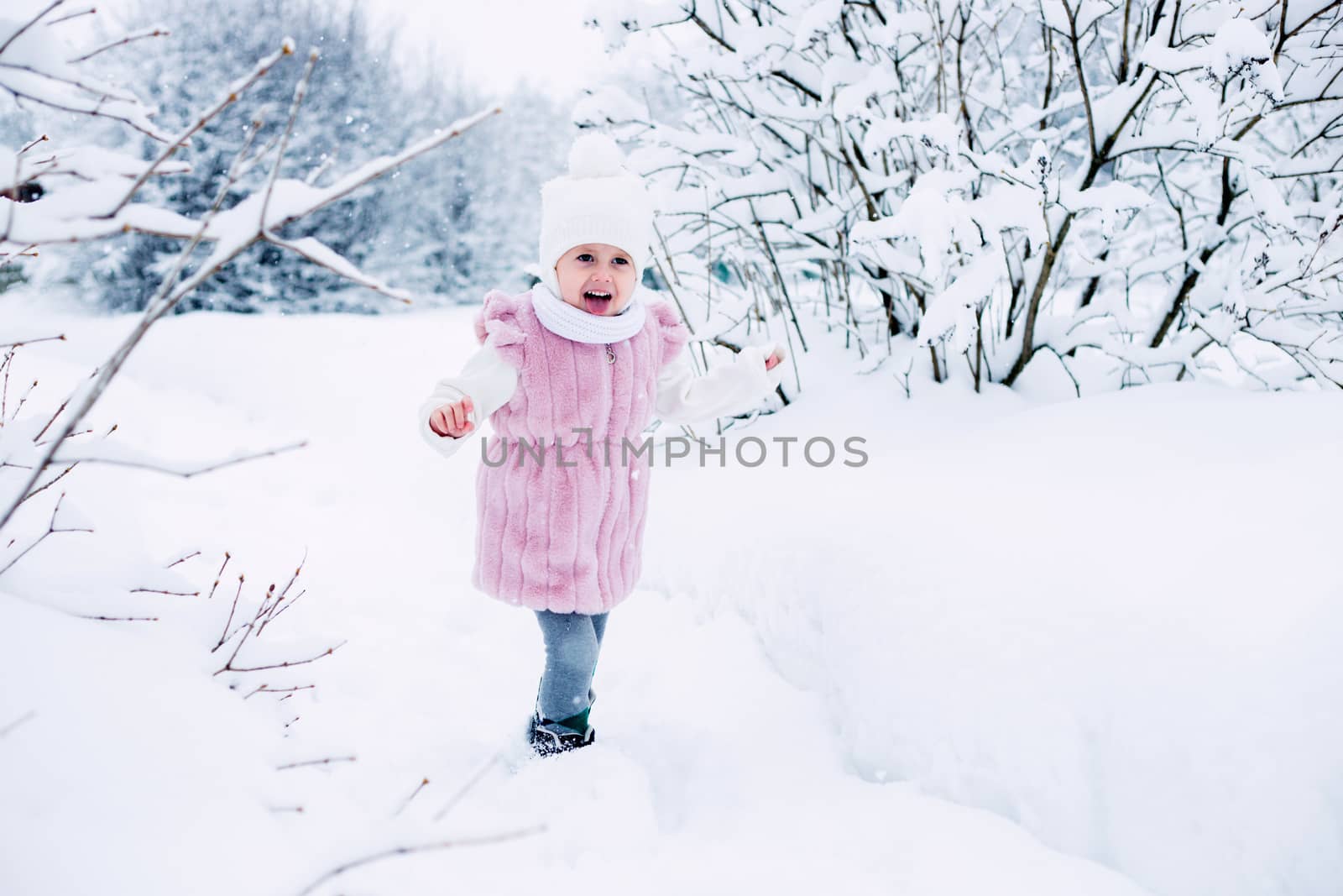 .A little girl in a pink fur coat stands in the middle of a snow-covered park on a cloudy winter day