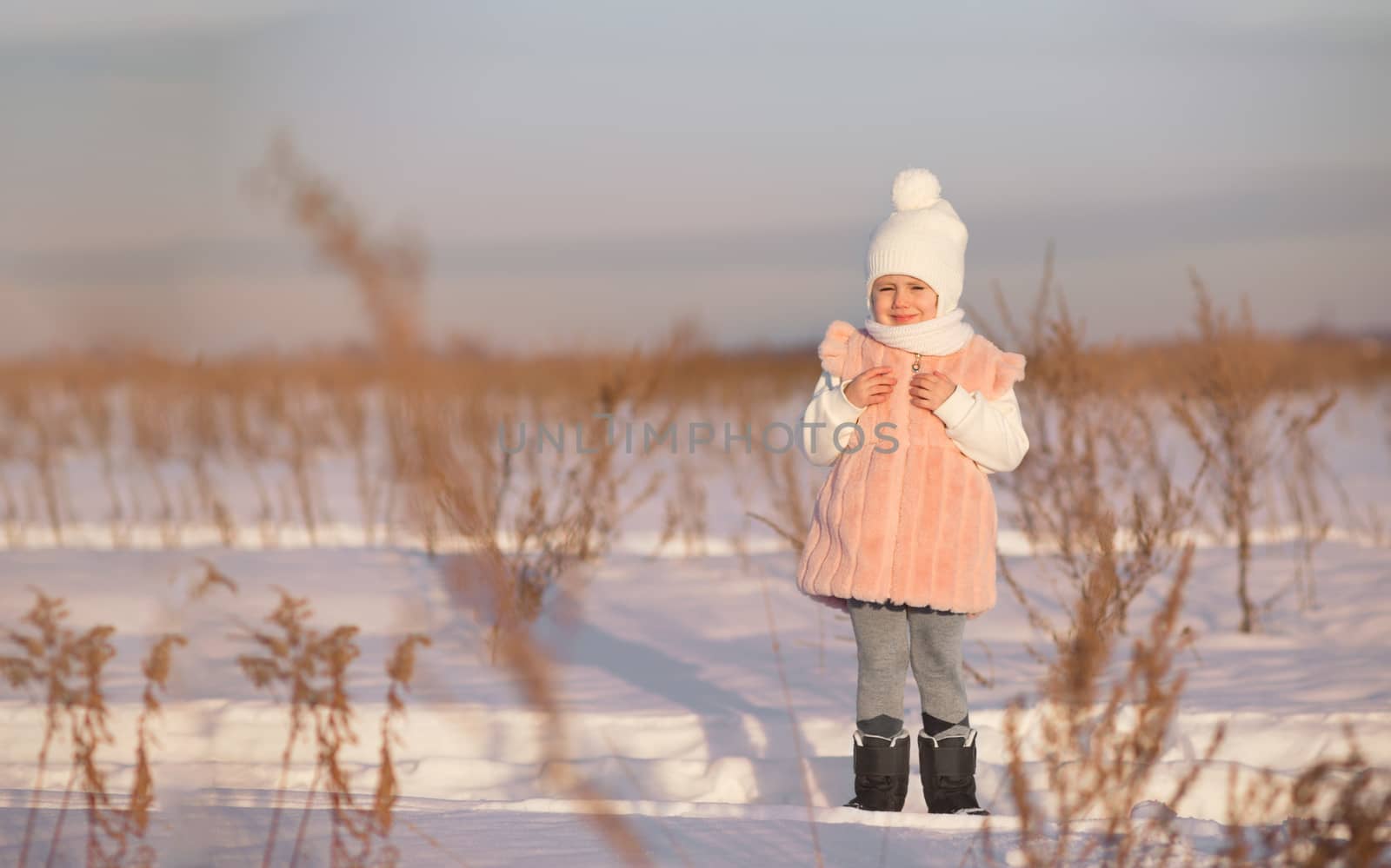 A little girl stands in the middle of a field in winter at sunset in the rays of the setting sun