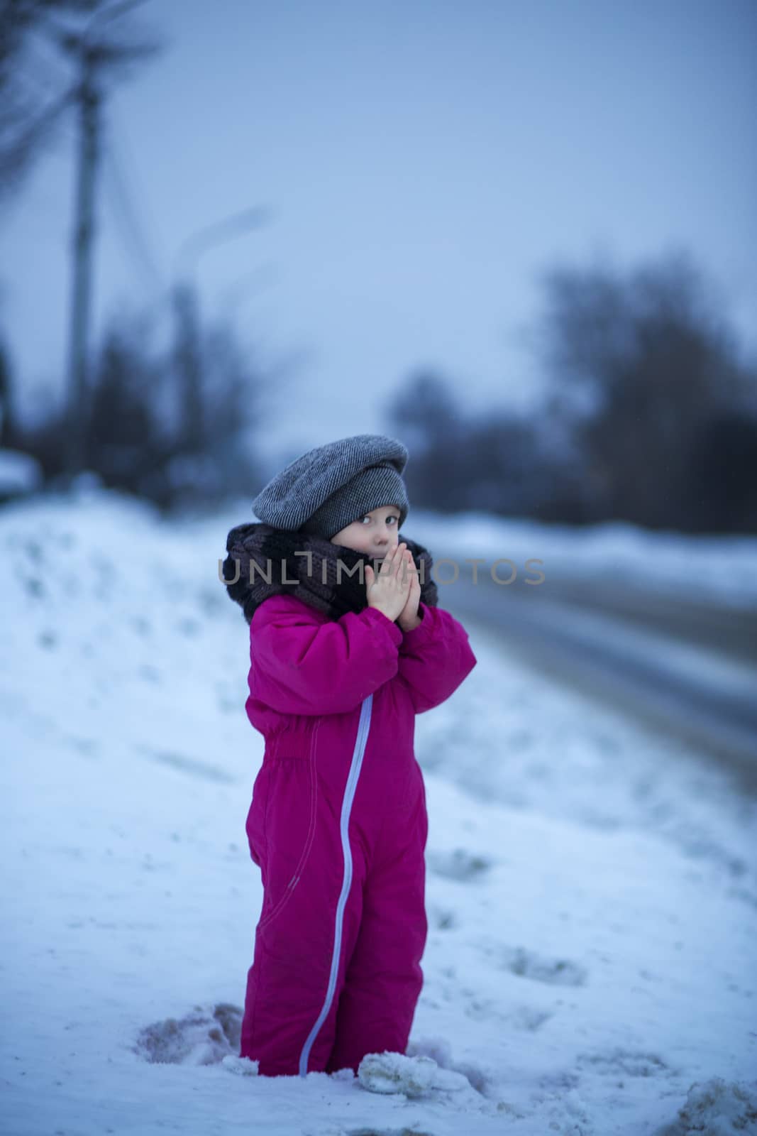 Little girl warms her hands with her breath on a winter evening near the road
