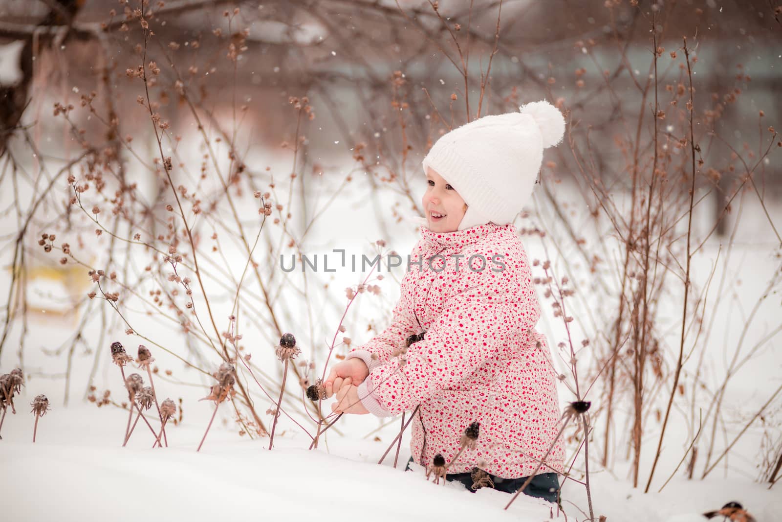A little girl sits in the snow and picks dried plants on sunny winter day