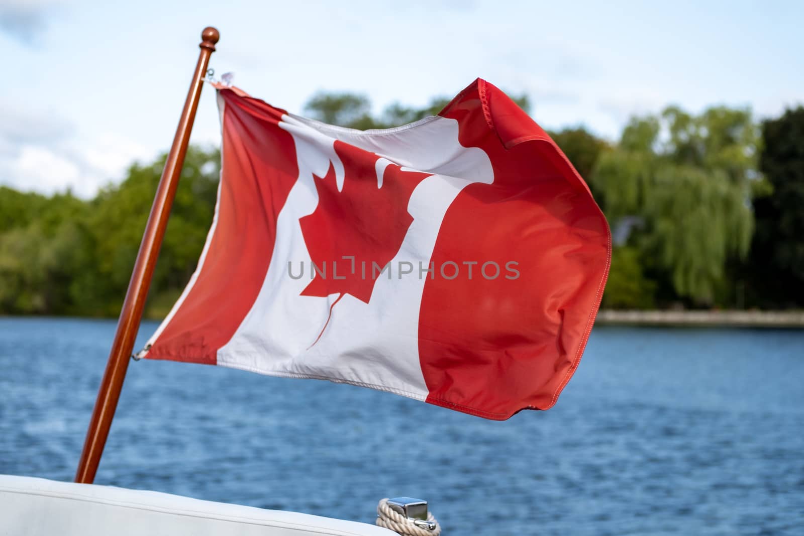 A Canadian flag flies in the wind at the stern, or back, of a small boat. Trees on a free. shore and the blue water of a river are seen behind it.