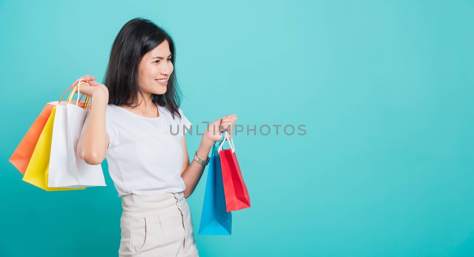 Portrait happy Asian beautiful young woman standing wear white t-shirt, She holding shopping bags multi color on hand and looking to space, shoot photo in a studio on a blue background