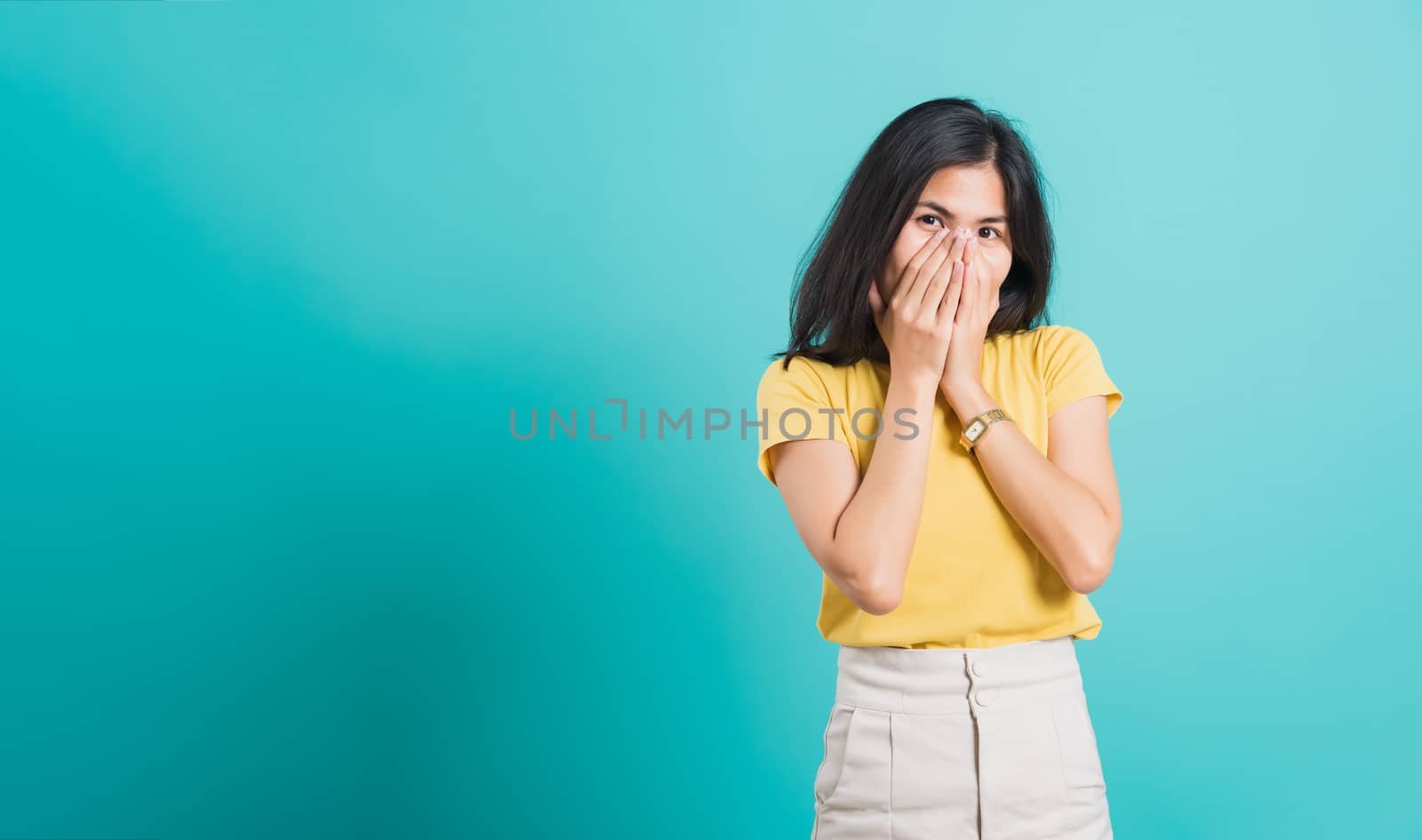 Portrait Asian beautiful happy young woman wears yellow t-shirt she happy excited laughs covering mouth with hands, shoot photo in a studio, on a blue background, with copy space