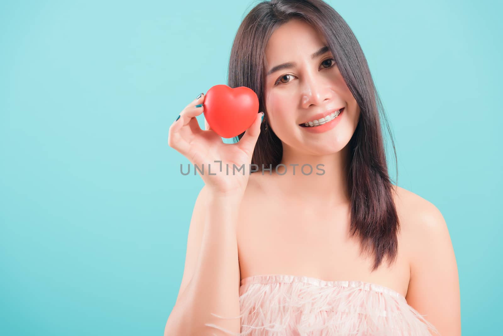 Asian happy portrait beautiful young woman standing smile holding red heart on hand near face on blue background with copy space for text