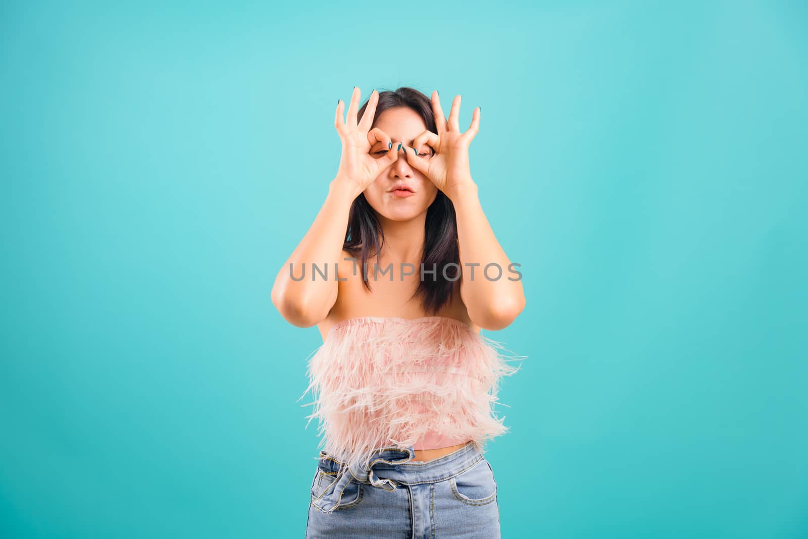 Smiling face asian beautiful woman her showing OK hand sign over her both eye and pucker up lips on blue background, with copy space for text