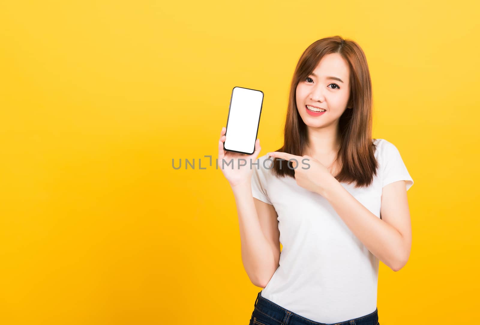 Asian happy portrait beautiful cute young woman smile standing wear t-shirt making finger pointing on smartphone blank screen looking to camera isolated, studio shot yellow background with copy space