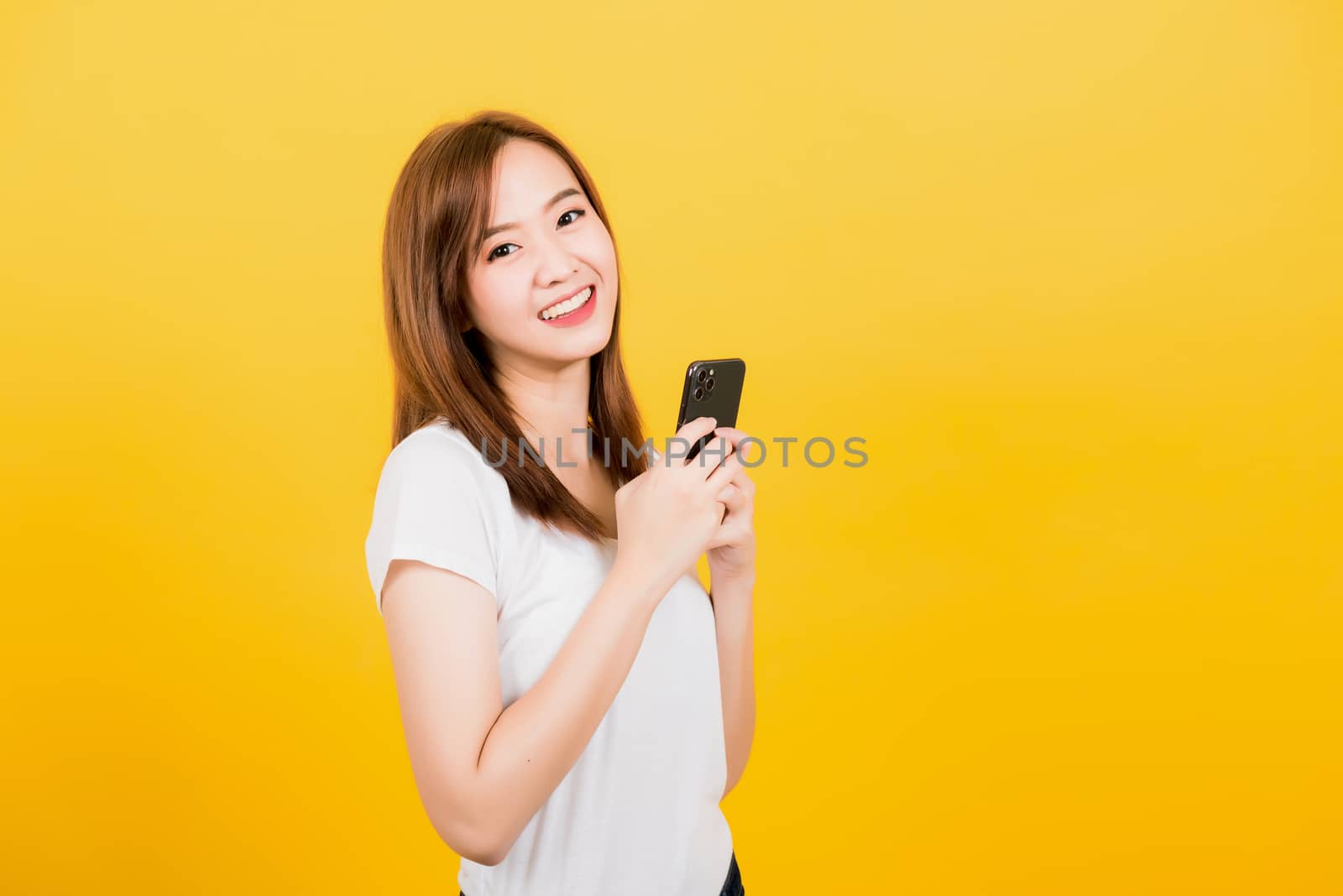 Asian happy portrait beautiful cute young woman teen smiling standing wear t-shirt using smart mobile phone looking to camera isolated, studio shot on yellow background with copy space