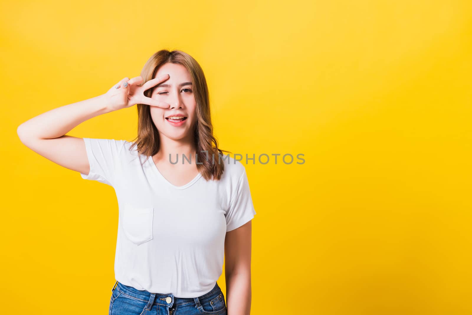 Asian Thai happy portrait beautiful cute young woman smile standing wear t-shirt showing finger making v-sign symbol near eye looking to camera, isolated studio shot yellow background with copy space