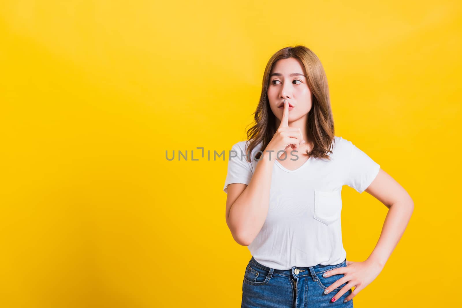 Asian Thai happy portrait beautiful cute young woman standing wear white t-shirt making finger on lips silent quiet gesture looking to side, studio shot isolated on yellow background with copy space