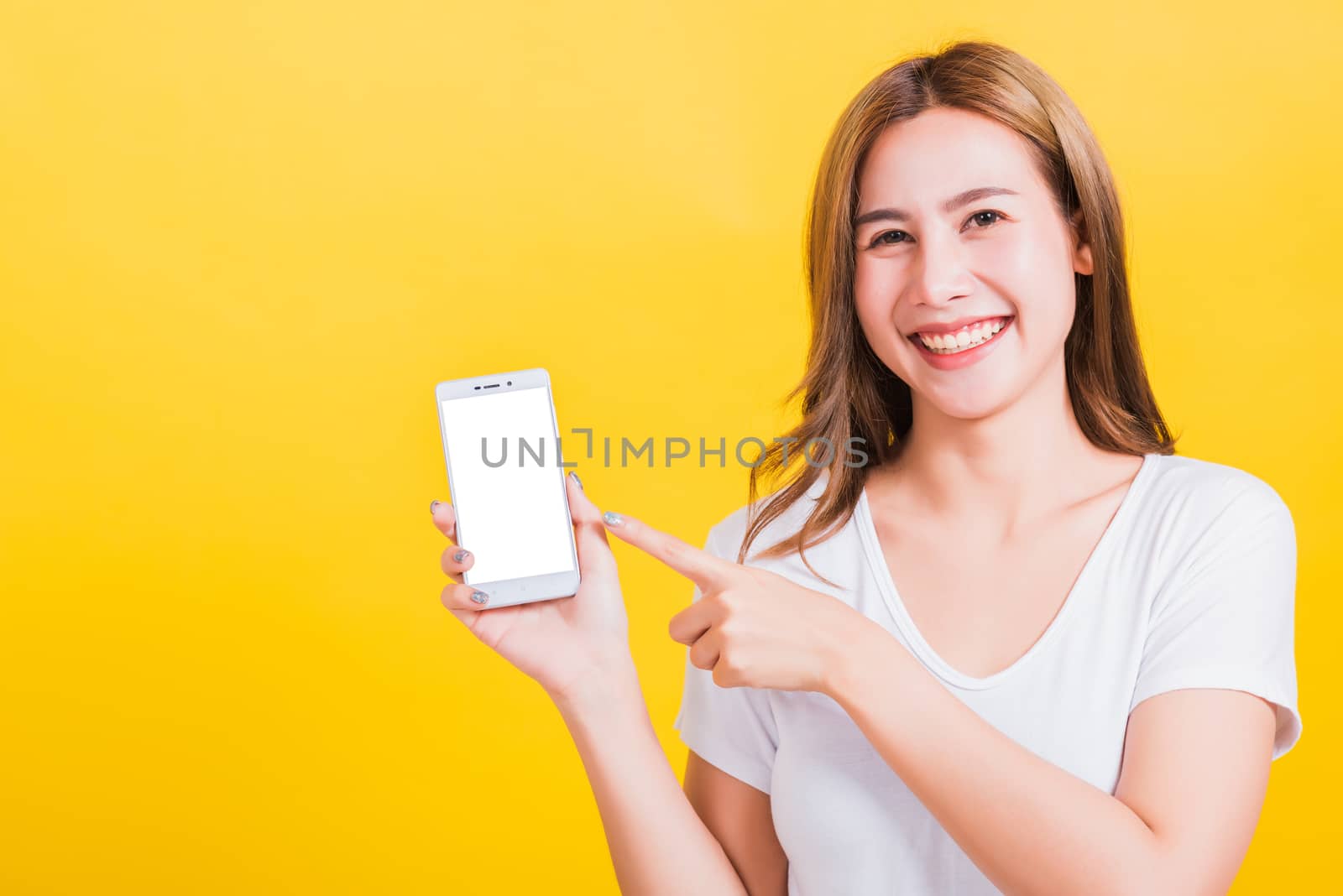 Asian Thai portrait happy beautiful young woman smile standing wear t-shirt making finger pointing on smartphone blank screen looking to camera isolated, studio shot yellow background with copy space