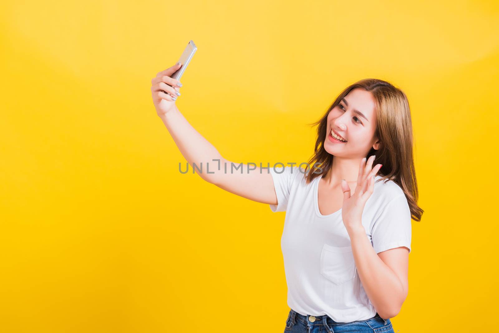 Asian Thai portrait happy beautiful young woman smiling making selfie photo or video call on smartphone she raise hand to say hello to smartphone, studio shot isolated yellow background and copy space