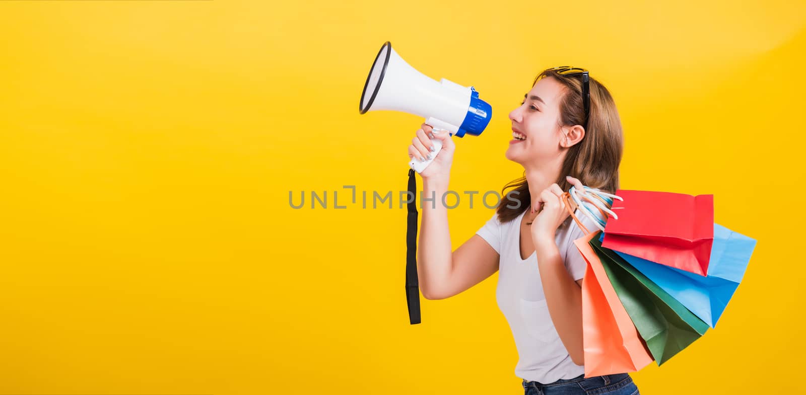 Portrait Asian Thai beautiful happy young woman smiling hold shopping bags multi-color and shouting in megaphone her looking to side away, studio shot isolated on yellow background, with copy space