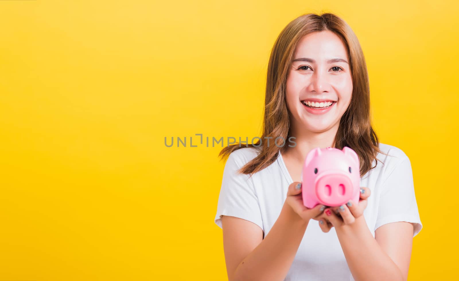 Portrait Asian Thai beautiful happy young woman smiling holding pink piggy bank with a lot of money and looking to camera, studio shot isolated on yellow background, with copy space