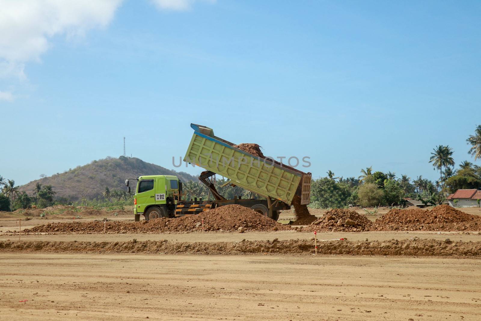 Green dump truck dumps its load of clay on construction site for road construction. Moto GP Mandalika racing circuit under construction, Lombok, Indonesia. Trucking industry, freight cargo transport.