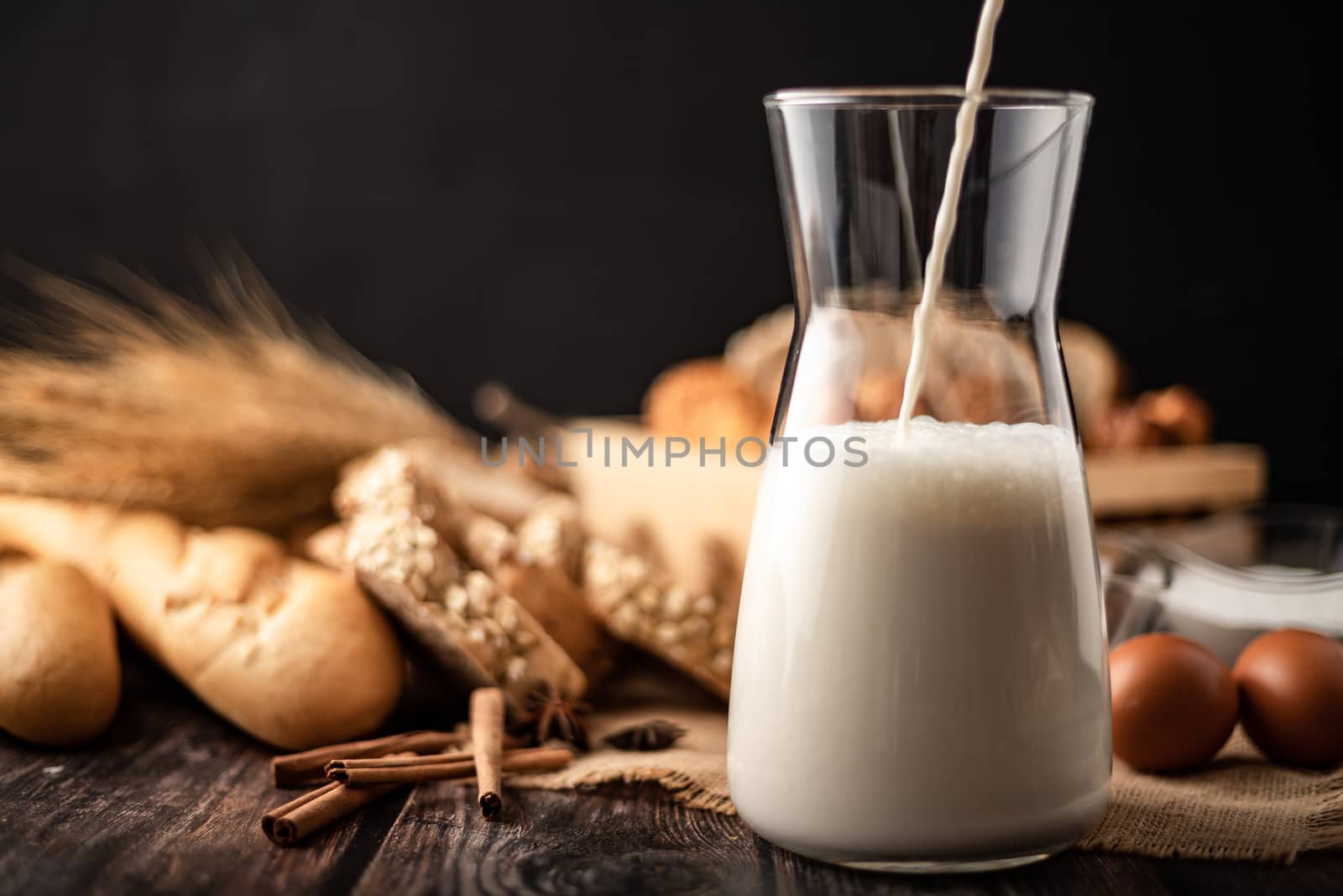 Pour the milk in a glass bottle placed on the sack. A lot of bread and eggs lay on a wooden table.