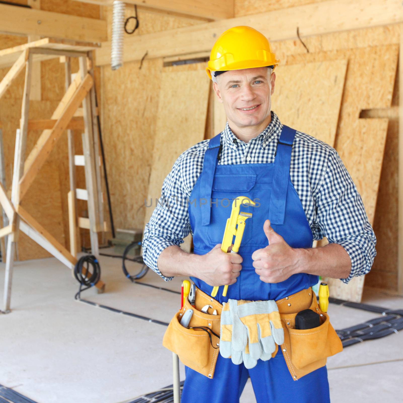 Happy male construction worker in blue uniform coveralls and yellow hardhat holding wrench showing thumb up and smiling