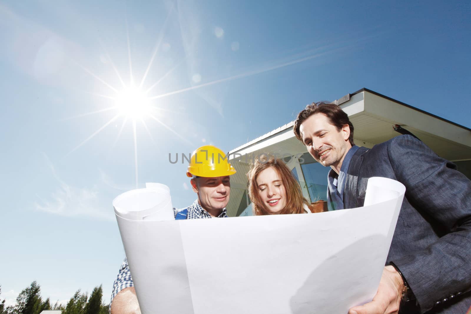 Worker shows construction plan to young couple outside house under construction sun in shining