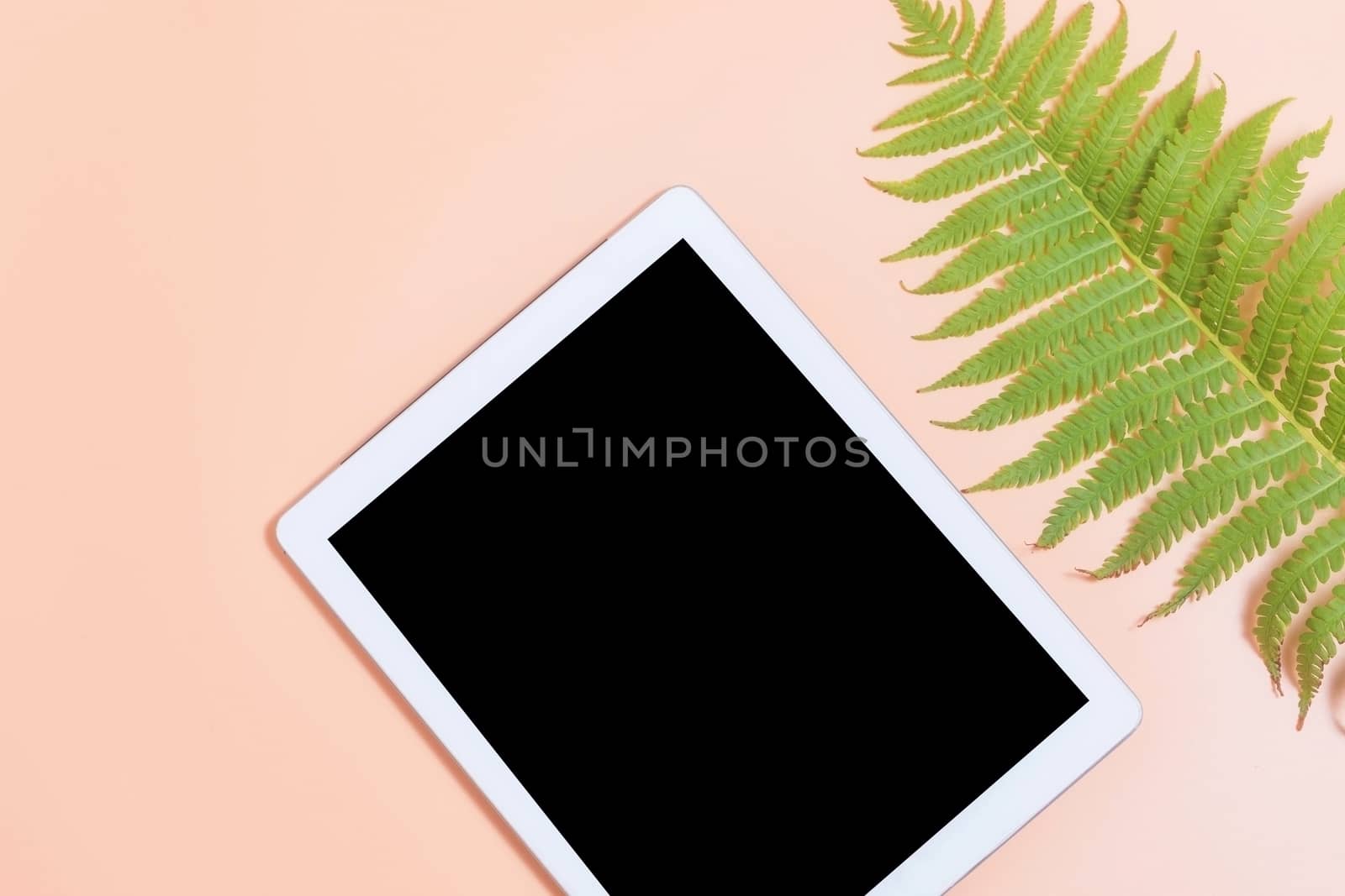 Tablet mockup on beige work desk surrounded with green leaf of fern. Free space beside for text. Flat lay composition.