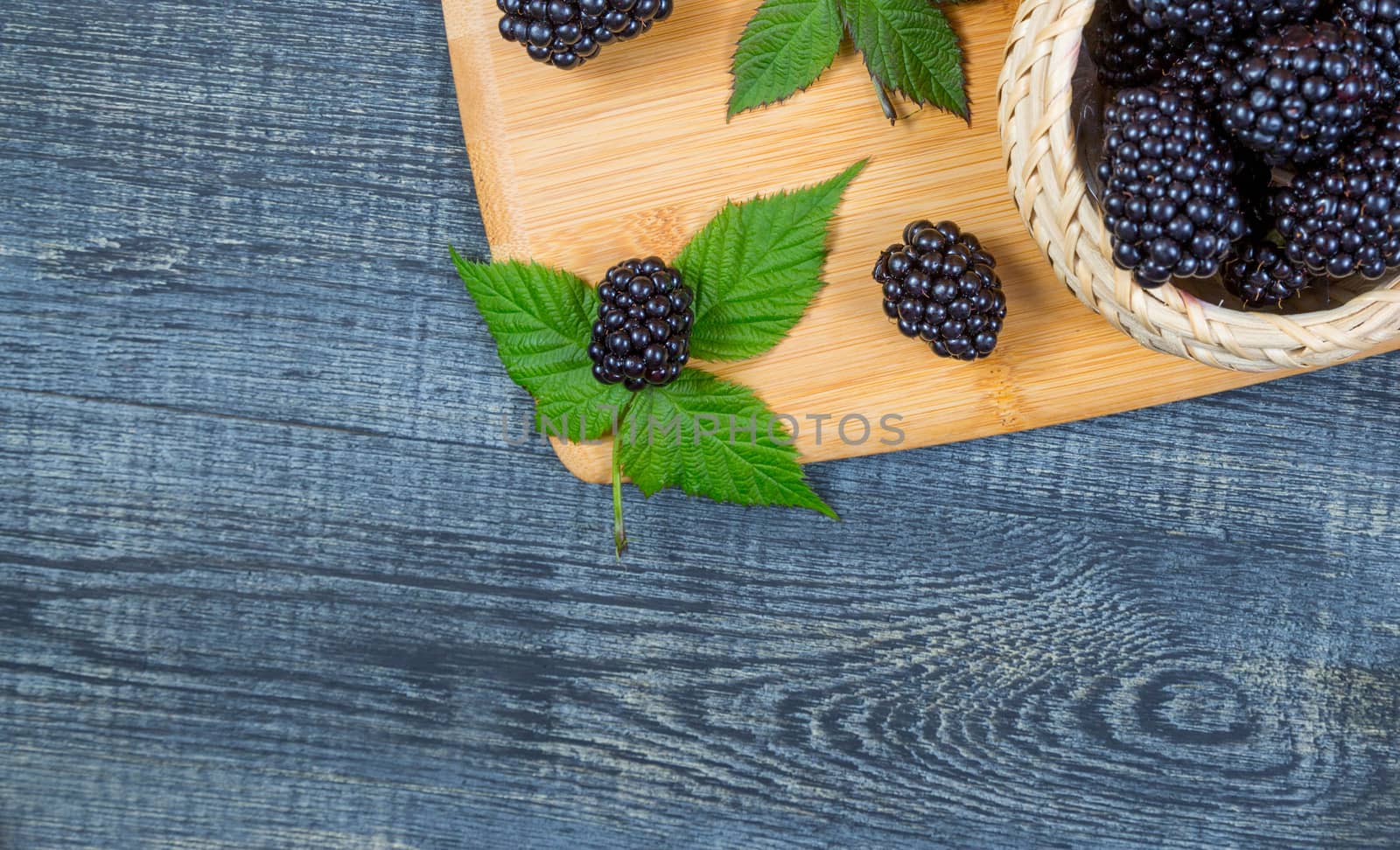 ripe blackberry with leaves on a wooden cutting board in a white ceramic bowl on dark blue wooden background. top view, place for text,