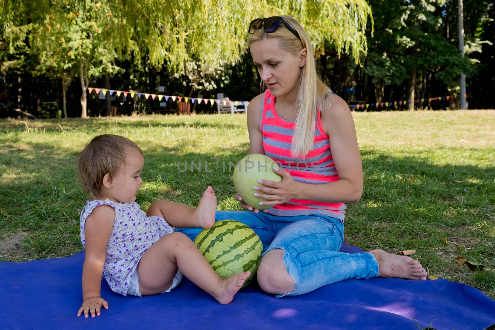 Young woman and little daughter sit in the park on the grass and play with watermelons. Family picnic.