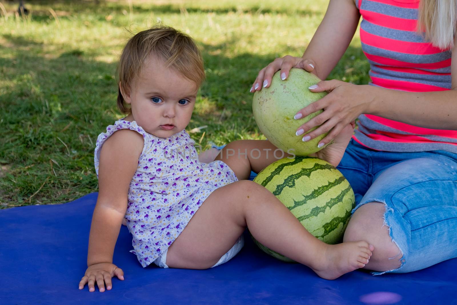 Young woman and little daughter sit in the park on the grass and play with watermelons. Family picnic. by leonik