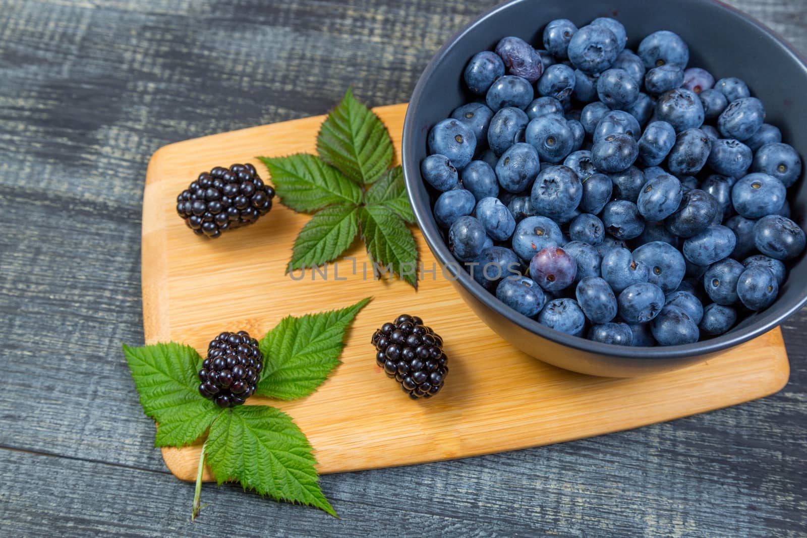 blueberry berry in dark gray ceramic bowl on wooden cutting board on dark blue wooden background.