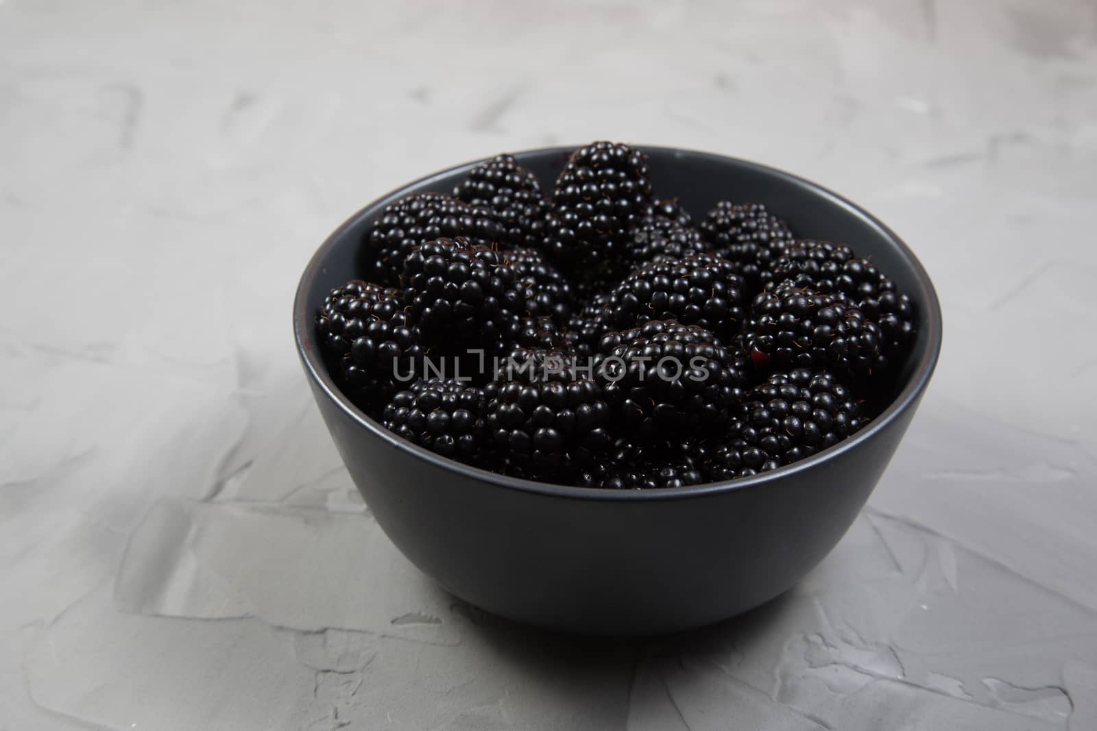 Ripe sweet blackberry in a gray ceramic bowl on gray concrete background.