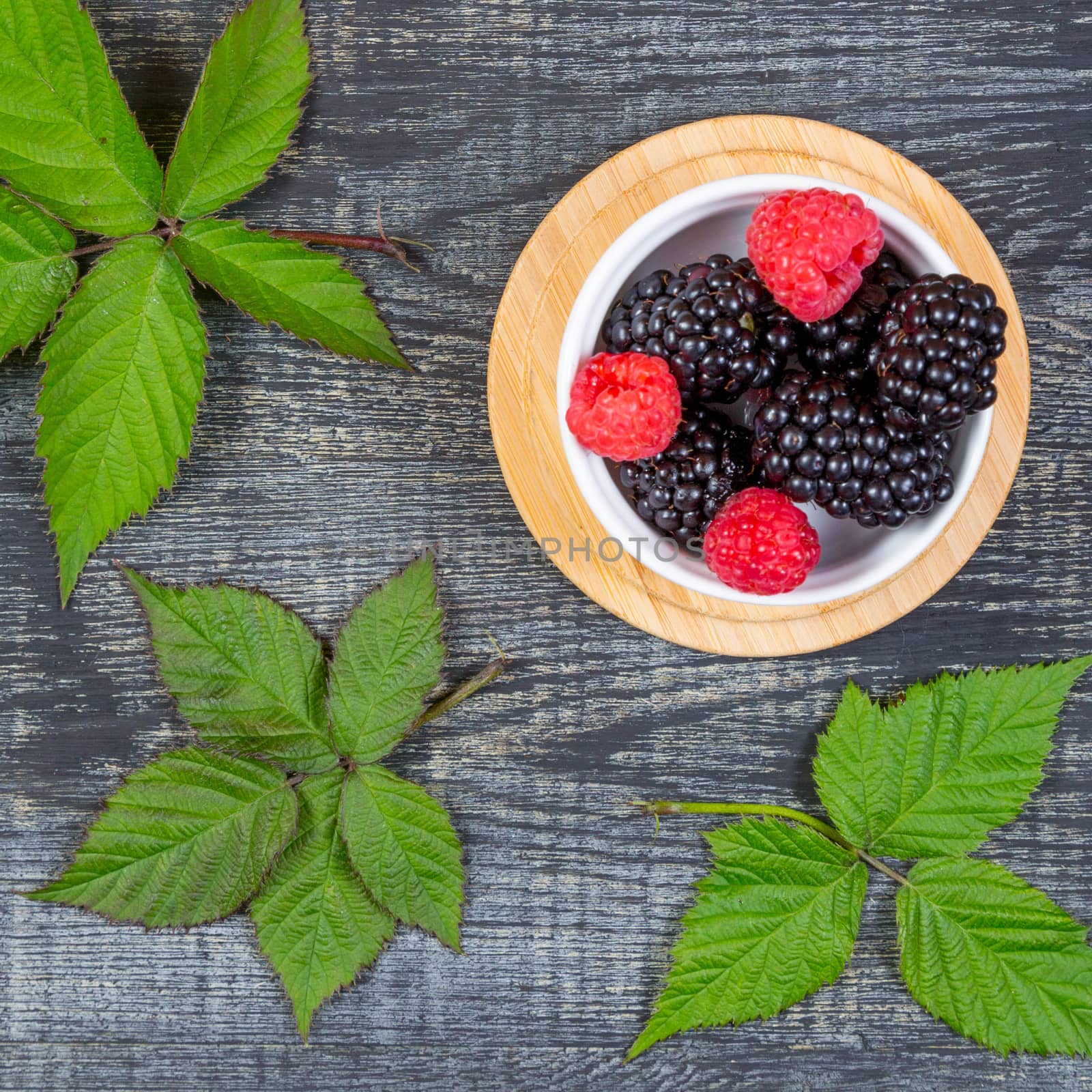 ripe blackberry with leaves on a wooden cutting board in a white ceramic bowl on dark blue wooden background. top view, 