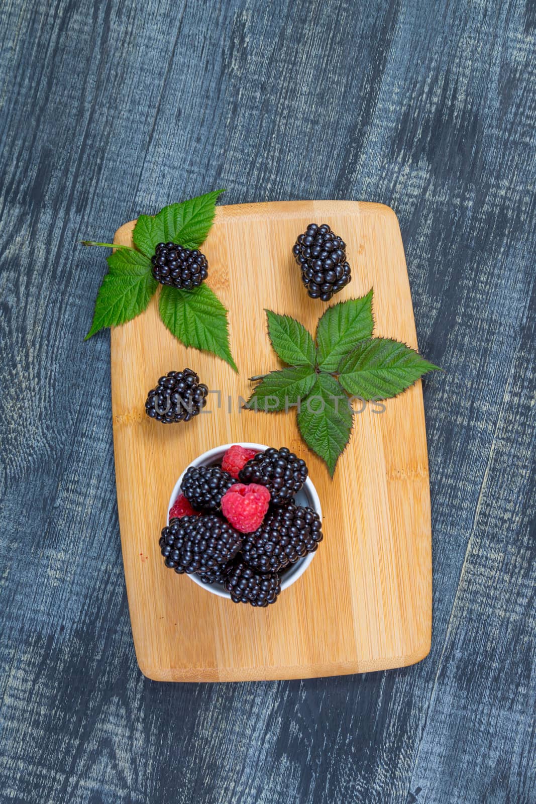 ripe blackberry with leaves on a wooden cutting board in a white ceramic bowl on dark blue wooden background. top view, place for text,