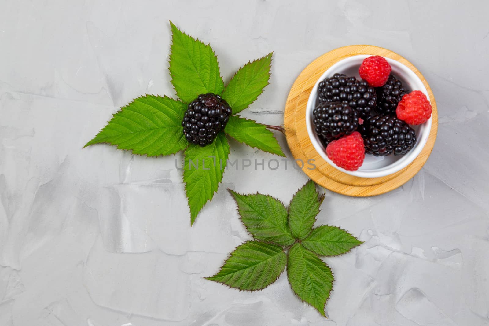 ripe blackberry with leaves on a wooden cutting board in a white ceramic plate on concrete background