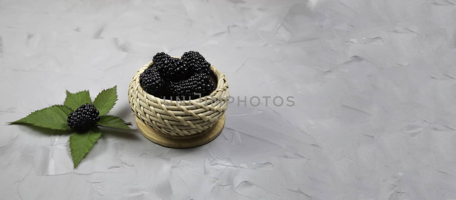 Ripe sweet blackberry in wooden wicker bowl on gray concrete background.