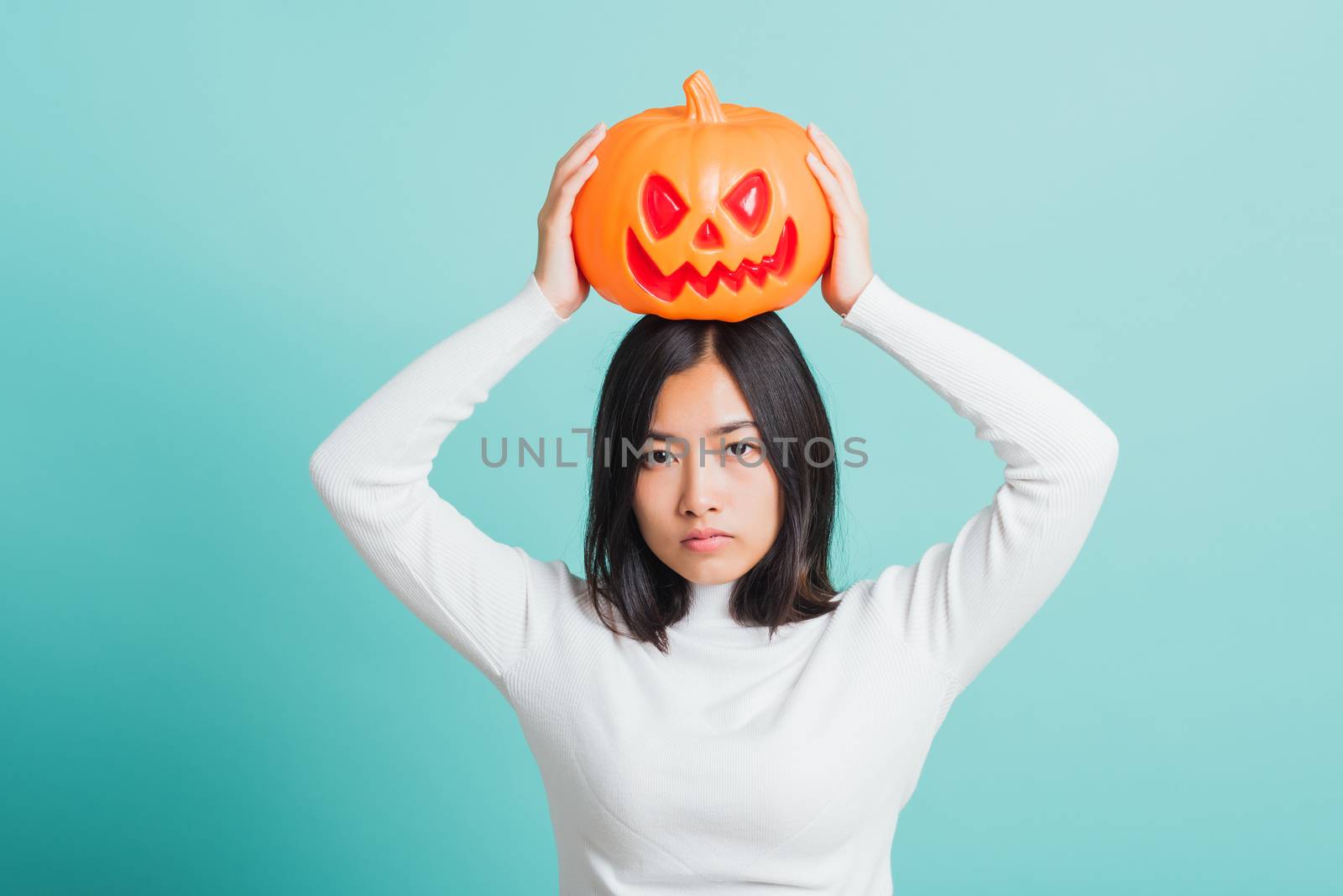 Portrait of Asian beautiful young woman holding orange model pumpkins and put it on the head, funny happy female with ghost pumpkins, studio shot isolated on blue background