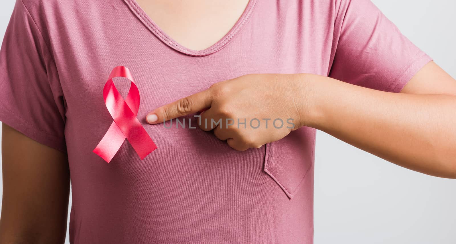 Breast cancer awareness healthcare and medicine concept. Close up Asian woman wear pink shirt pointing finger to pink breast cancer awareness ribbon, studio shot isolated on white background