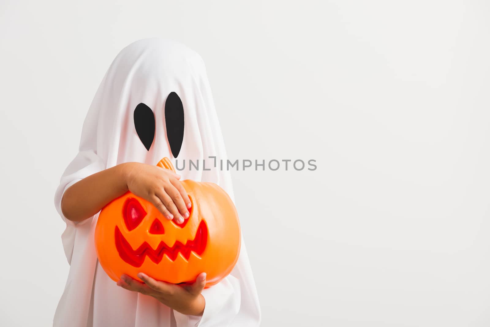 Funny Halloween Kid Concept, little cute child with white dressed costume halloween ghost scary he holding orange pumpkin ghost on hand, studio shot isolated on white background