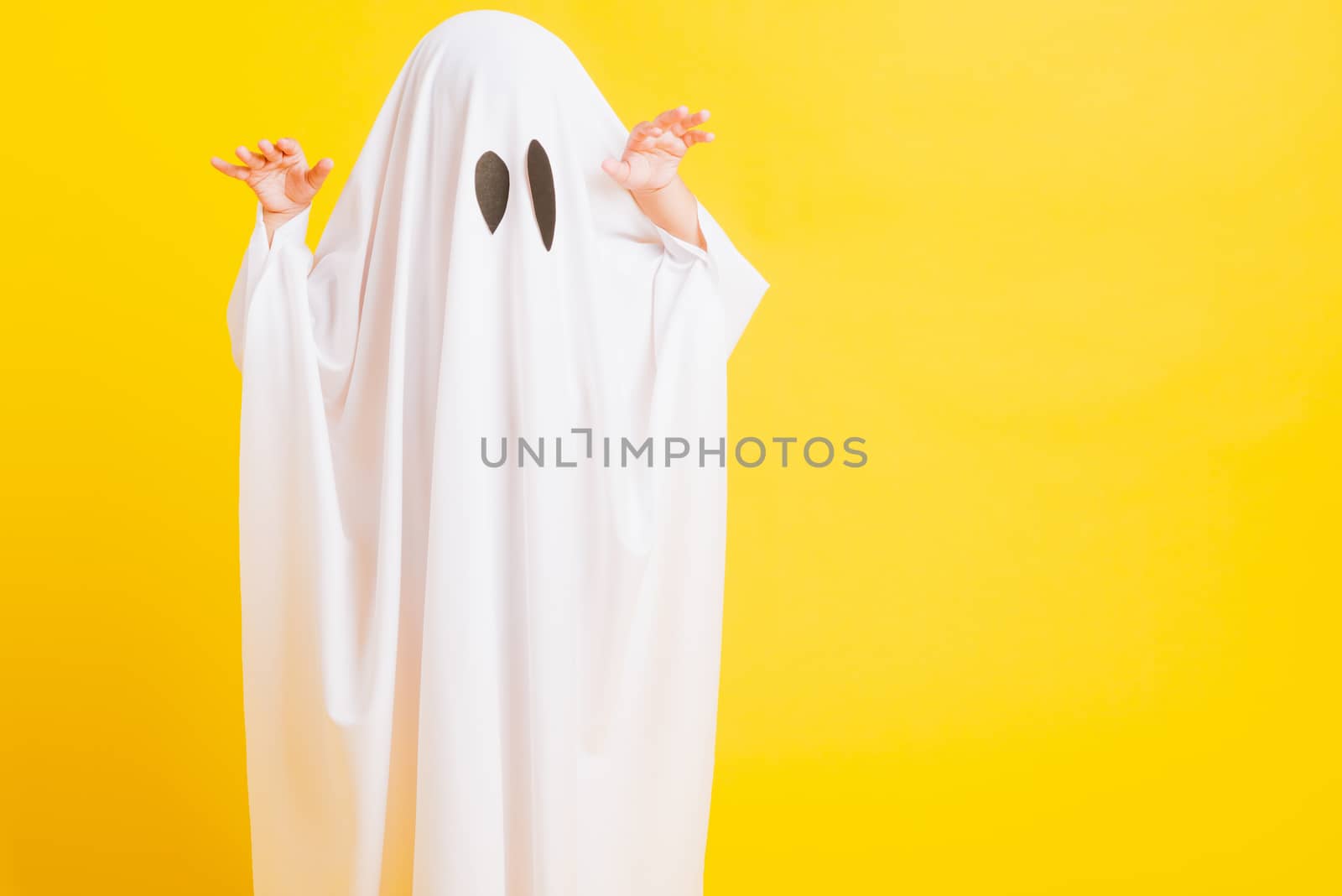 Funny Halloween Kid Concept, Closeup a little cute child with white dressed costume halloween ghost scary, studio shot isolated on yellow background