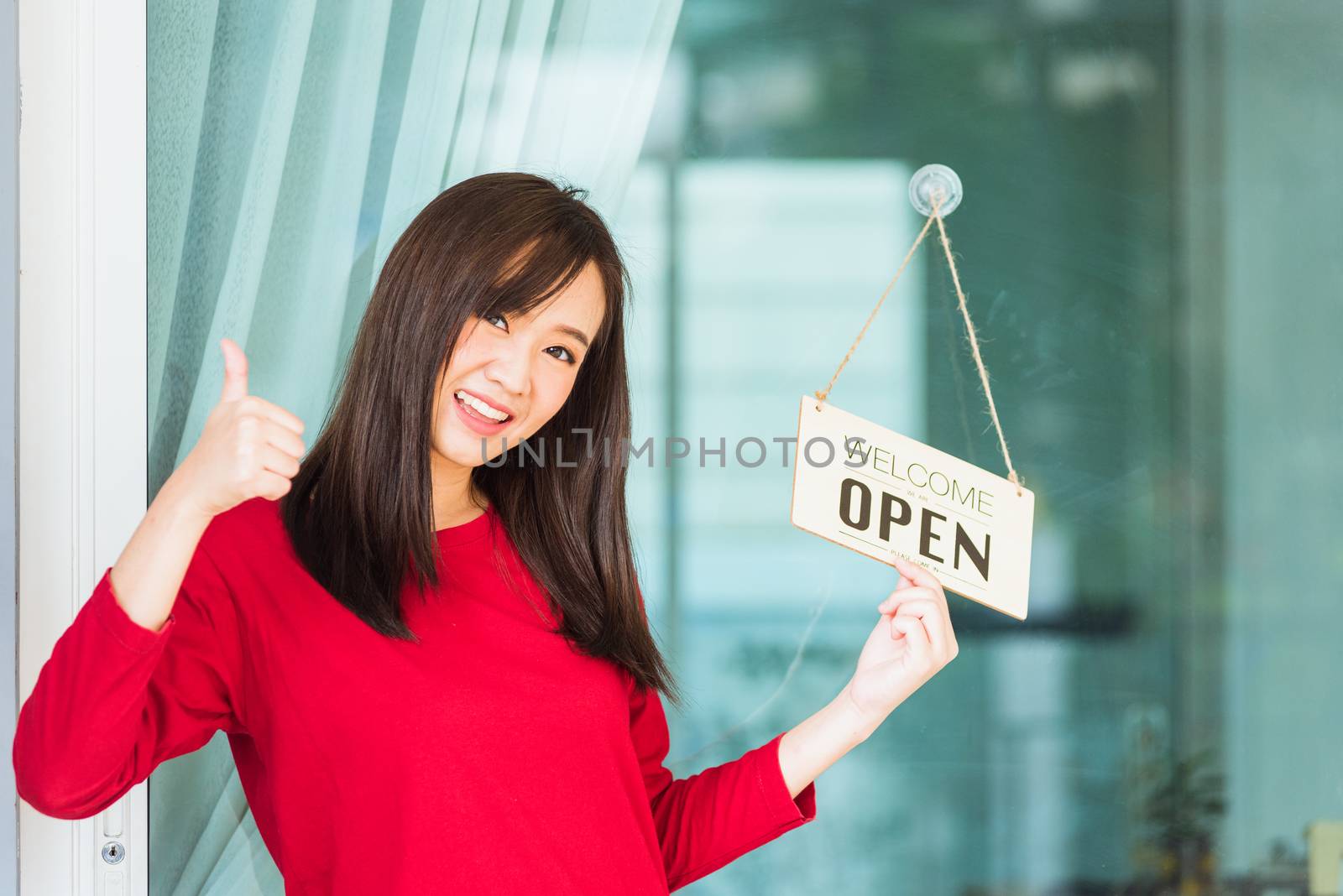 Asian young woman smile show finger thumb up for good sign she notice sign wood board label "WELCOME OPEN" hang through glass door front shop, Business turning open after coronavirus pandemic disease