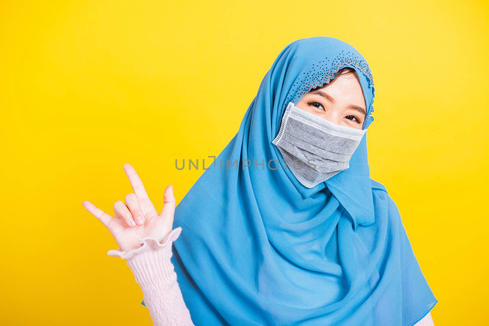Asian Muslim Arab, Portrait of happy beautiful young woman Islam religious wear veil hijab and face mask protect quarantines disease coronavirus show hand finger "I LOVE YOU" sign isolated yellow